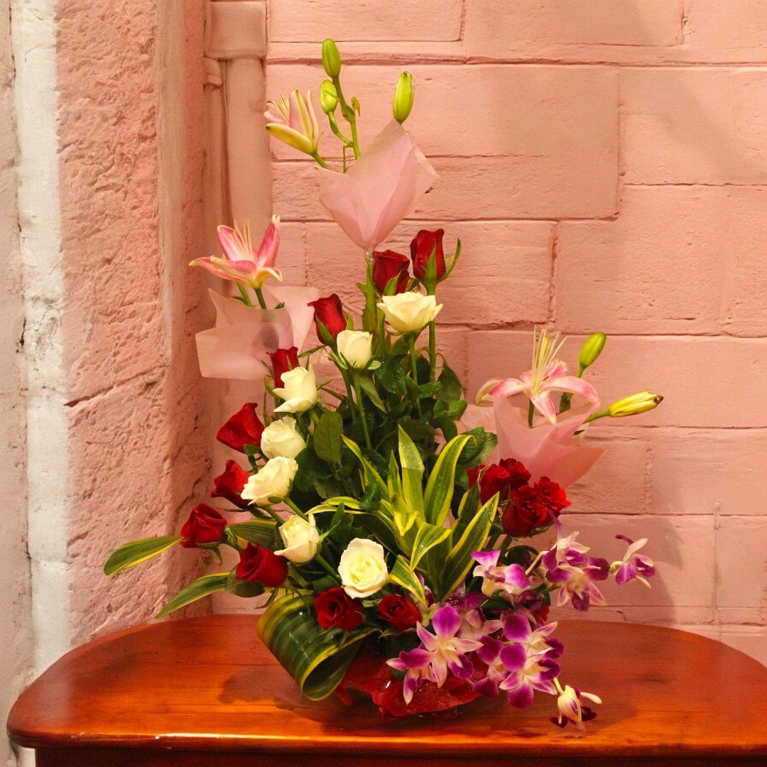 A beautiful arrangement of red and white roses, pink lilies, and orchids by BetterGiftFlowers placed on a wooden table against a pink brick wall.