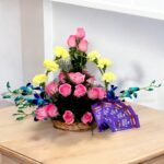 A basket of pink roses, yellow carnations, blue orchids, and Dairy Milk chocolates by BetterGiftFlowers.