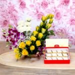 BetterGiftFlowers Mix Flower Basket N Kaju Katli with fresh yellow roses, orchids, and white carnations alongside a box of Haldiram's Kaju Katli.